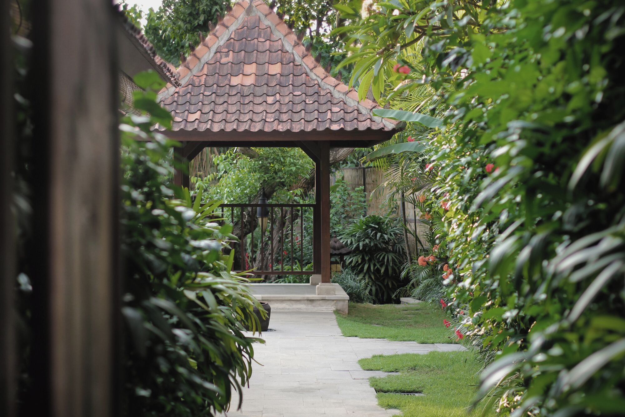 A garden pathway leading to a gazebo with a tiled roof, surrounded by lush greenery and flowering plants.