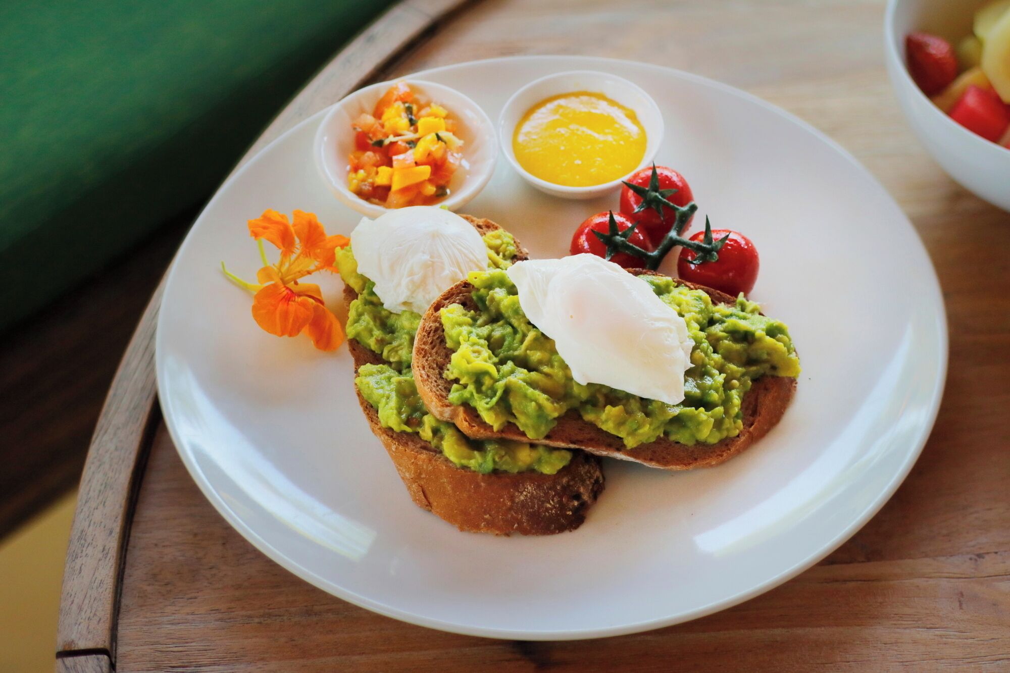 A plate with toast topped with avocado and poached egg, accompanied by a small bowl of salsa, a bowl of hollandaise sauce, and a cherry tomato.
