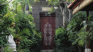 Une porte en bois traditionnelle, ornée de sculptures complexes, entourée d’une végétation luxuriante et bordée d’un chemin de pierre menant à un bâtiment dont le toit est recouvert de tuiles.