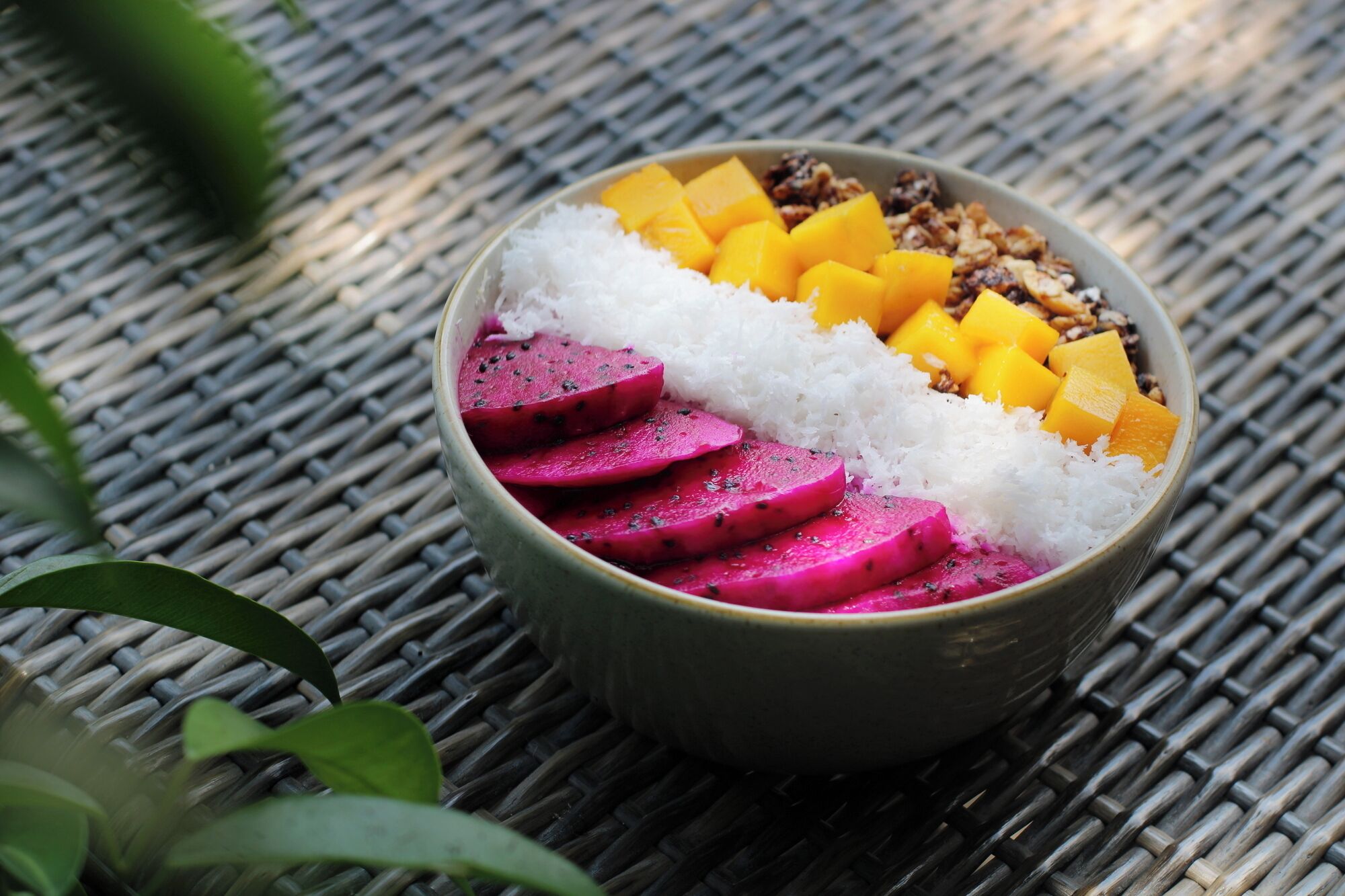 A bowl of food with white rice, yellow mango chunks, and pink dragon fruit slices on a woven mat.