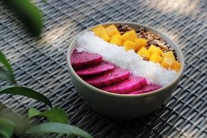 Un tazón de comida con arroz blanco, trozos de mango amarillo y láminas de pitahaya rosa sobre un tapete tejido.