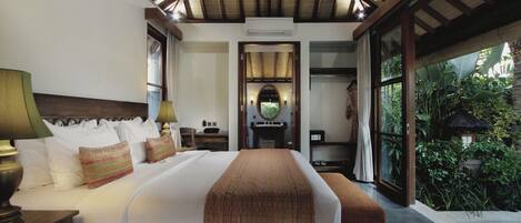 A spacious bedroom with a large bed, a nightstand, a desk, and a view of a lush garden through a glass door.