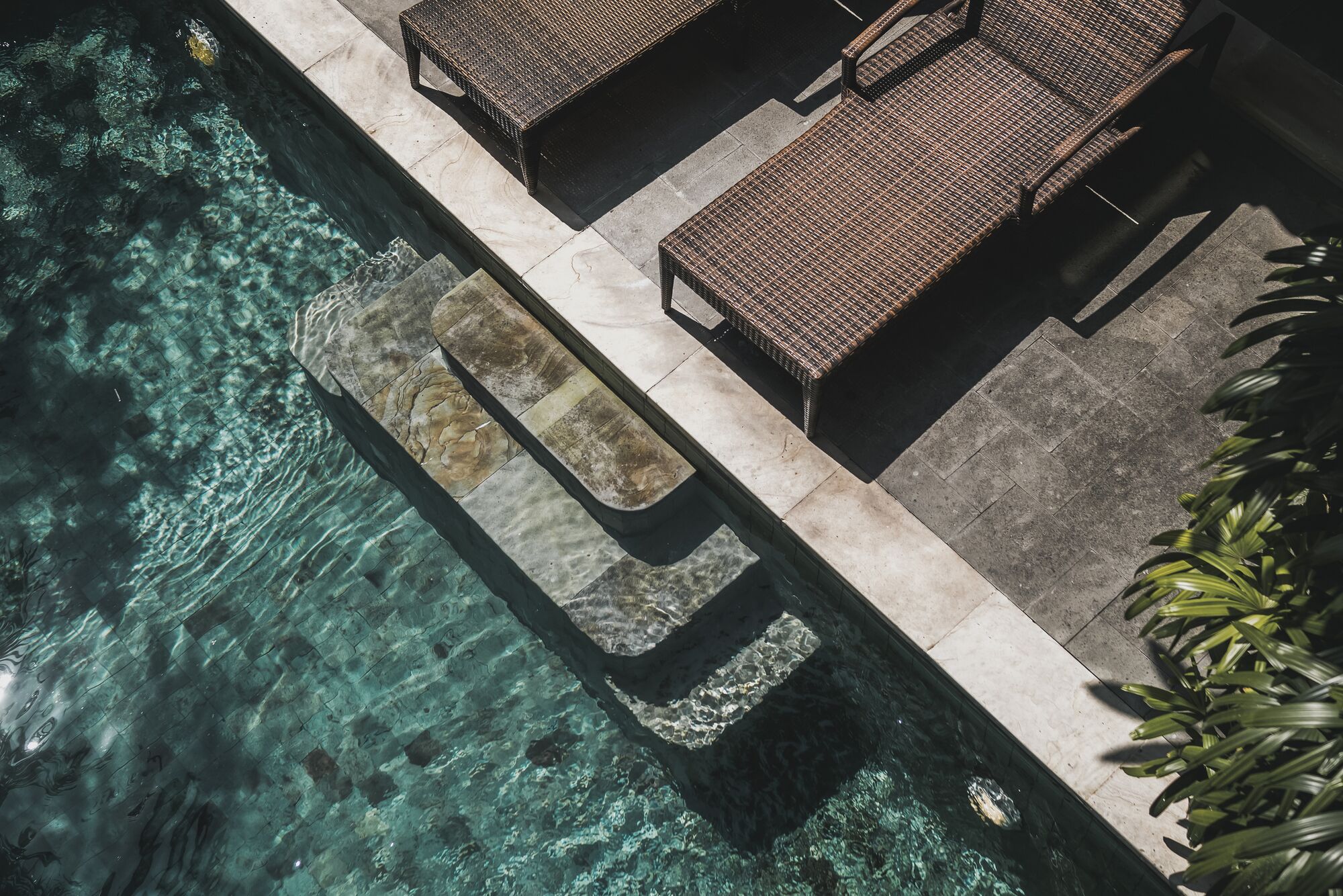 An aerial view of a pool area with clear water, lounge chairs, and a stone pathway.