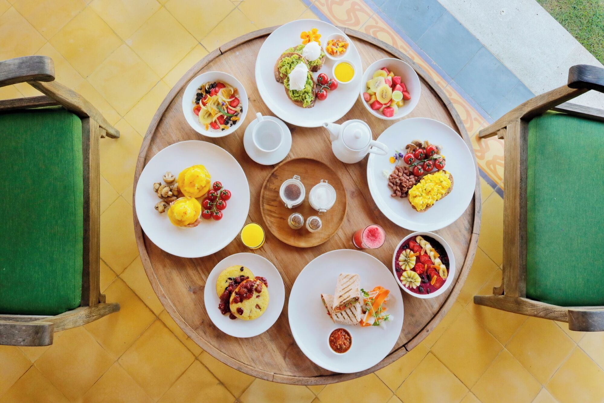 A round wooden table with various breakfast dishes and condiments, surrounded by green cushioned chairs.