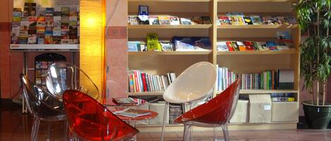 A library with bookshelves, a table with magazines, and clear plastic chairs.