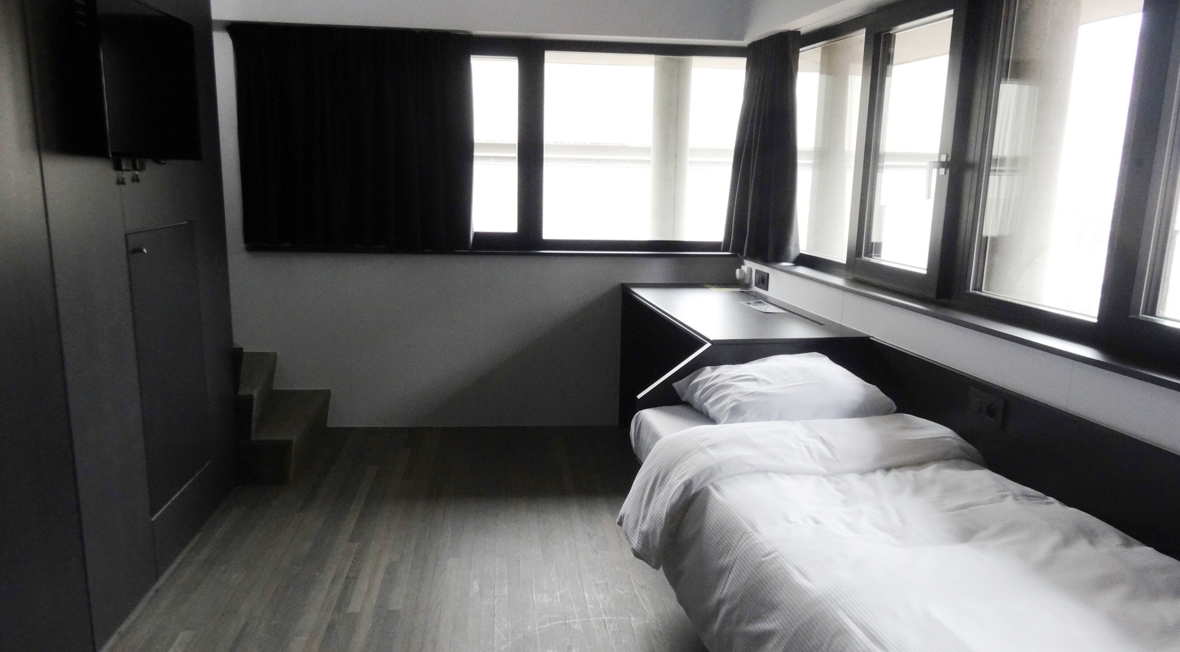 Quarto de hotel com cama, escrivaninha e televisão fixada na parede.