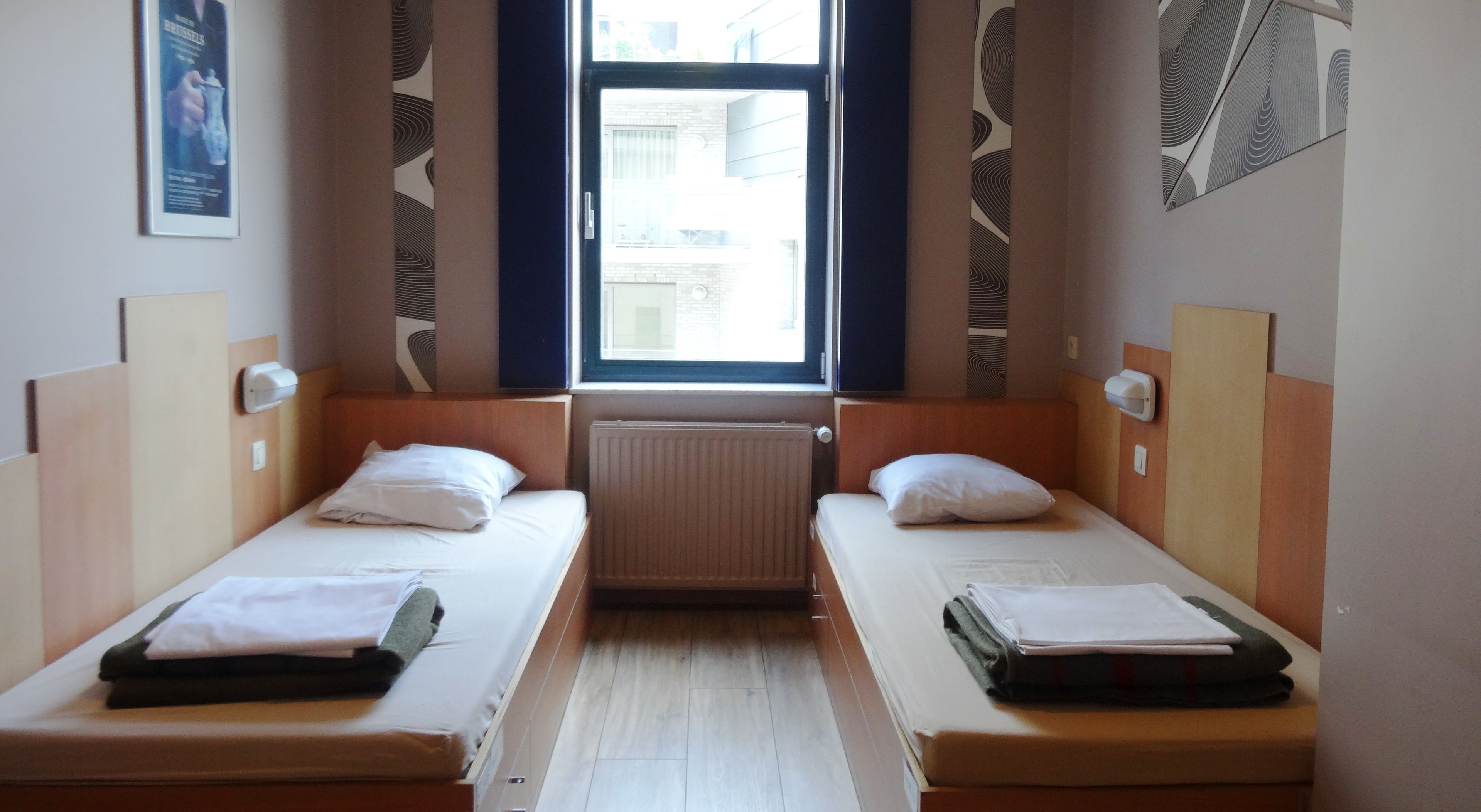 Habitación de hotel con dos camas individuales, una ventana, un radiador y un secador de manos instalado en la pared.