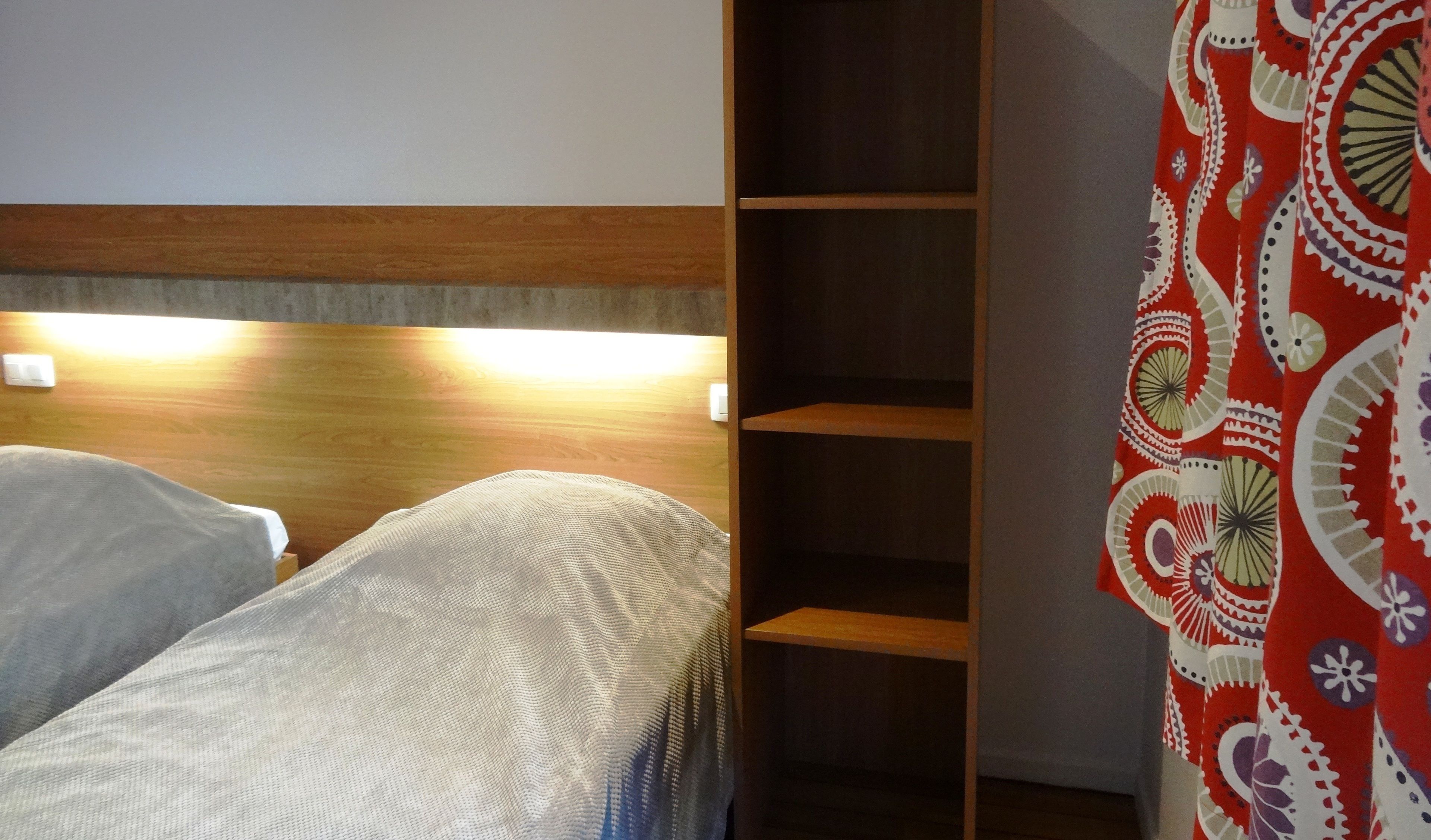 A bedroom with a wooden headboard, a bed with a white and gray patterned bedspread, and a red and white patterned curtain.