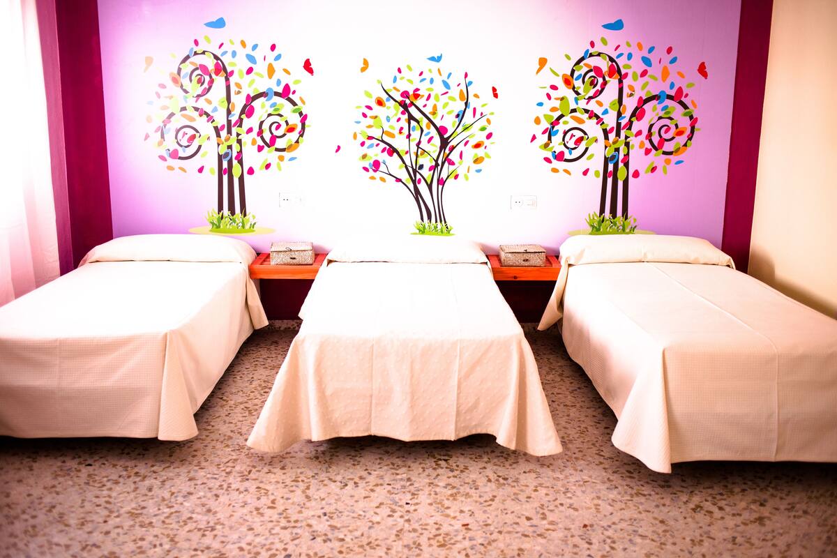 A hotel room with three single beds, a small table, and a wall with colorful tree decals.