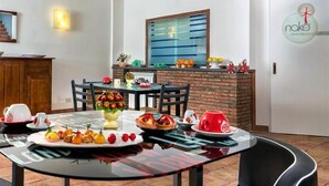 A dining area with a glass table set for a meal, a brick accent wall, and a kitchen area with a window and a logo on the door. - Nake Residenza Artistica (Sant'Alfio)