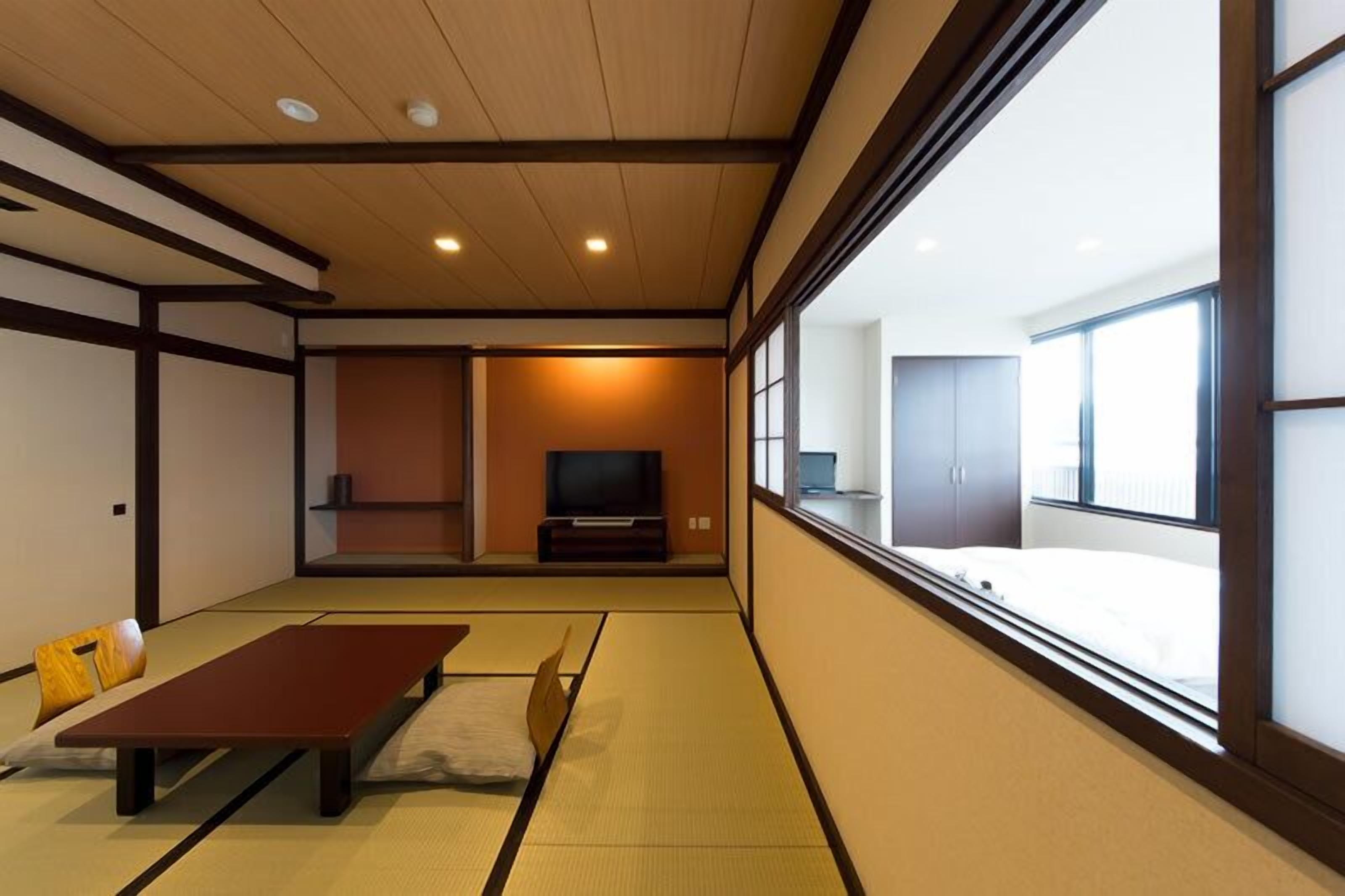 Una habitación japonesa tradicional con suelo de tatami, una mesa baja, un televisor y un ventanal que ofrece vistas al exterior.