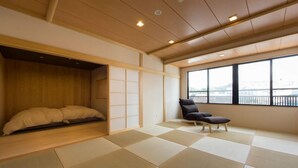 A room with tatami flooring, a futon bed, a sliding shoji screen, a black chair, and a view of a bridge through large windows. - Hotel Kikyou (Kaga)