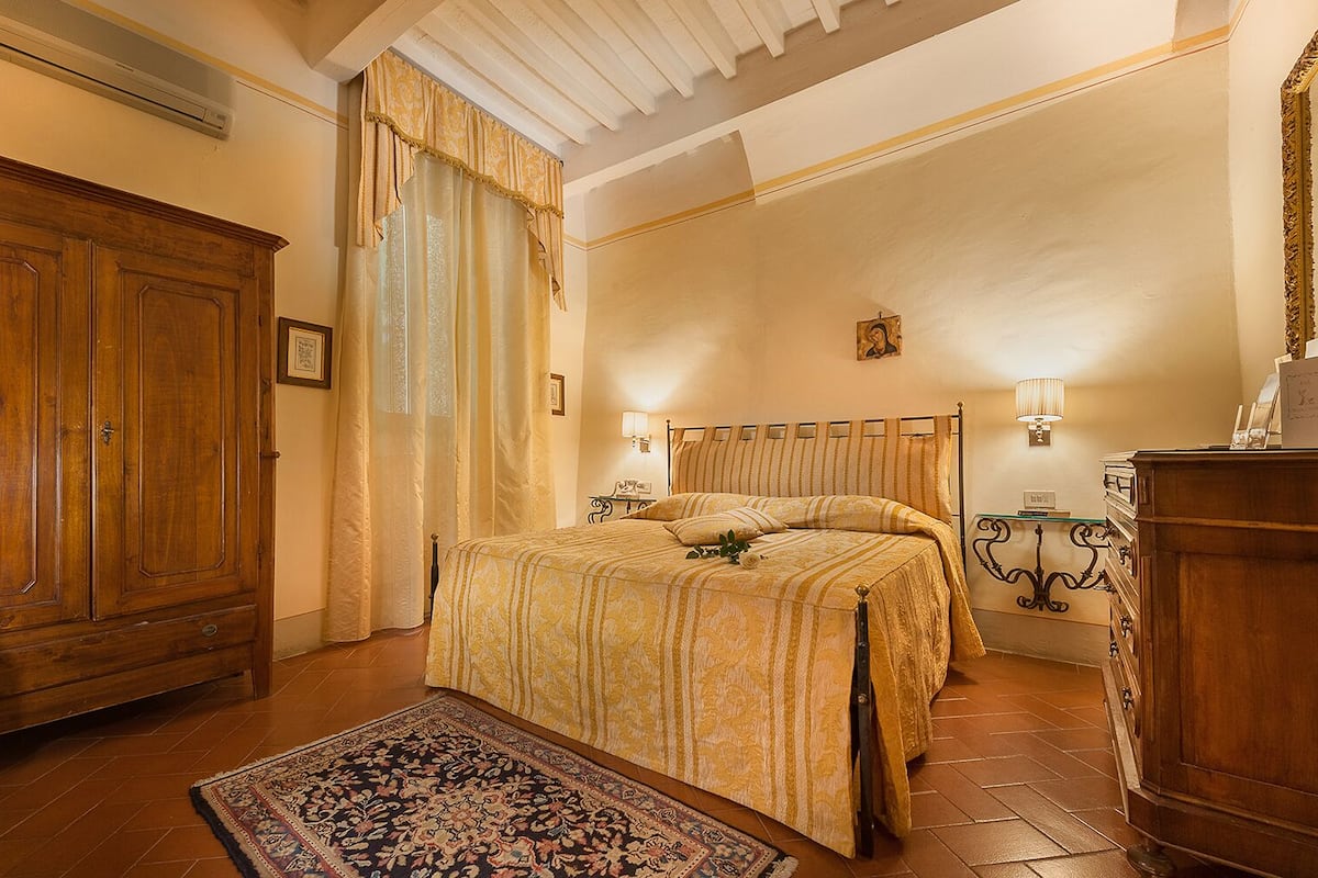 A bedroom with a large wooden wardrobe, a bed with a patterned bedspread, a decorative rug, and a nightstand with a lamp.