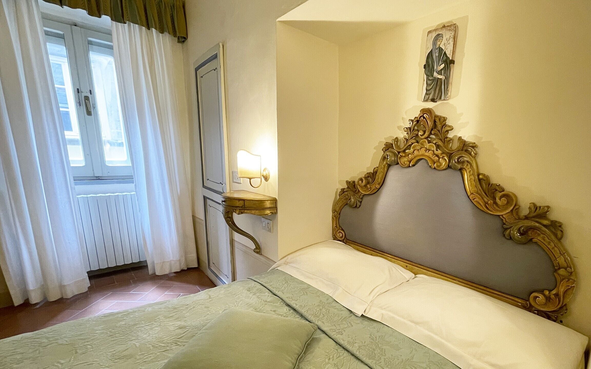 Una habitación con cama, ventana con cortinas, lámpara de pared y un cuadro pequeño.