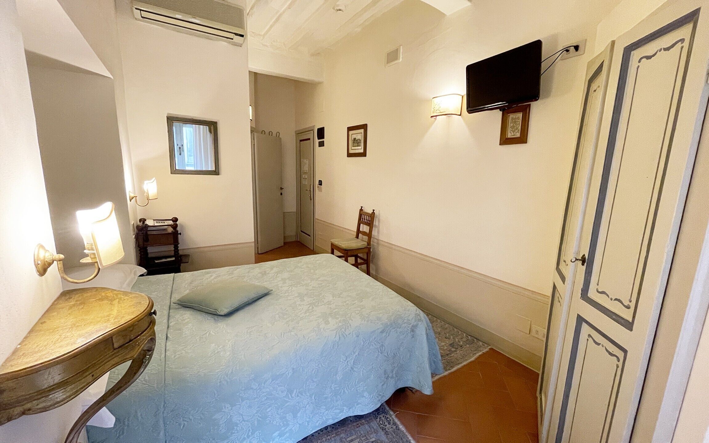 Un dormitorio con cama, mesita de noche, una silla, televisor en la pared y una puerta.