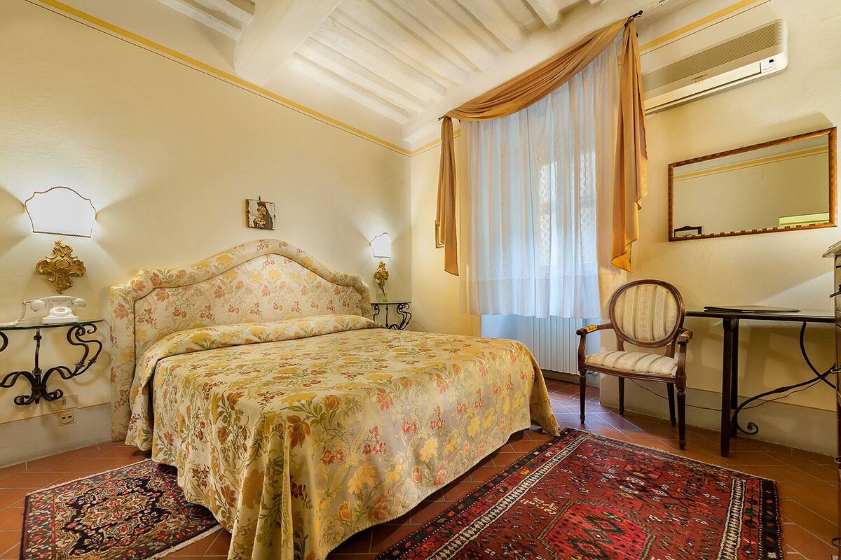 A bedroom with a floral bedspread, a wooden chair, a desk, and a mirror.