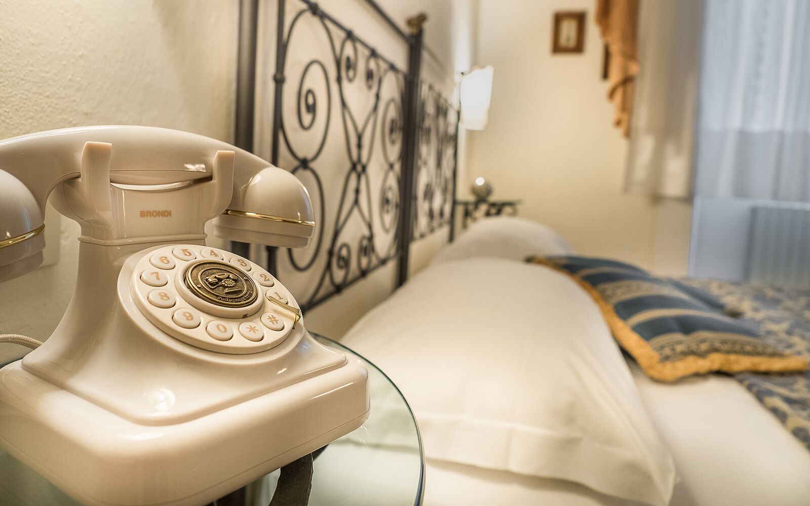 Ein altmodisches weißes Wählscheibentelefon auf einem Nachttisch in einem Hotelzimmer mit einem Bett, einem dekorativen Metall-Kopfteil und einem kleinen Beistelltischchen mit einer Lampe.