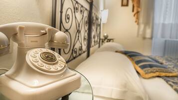 Ein altmodisches weißes Wählscheibentelefon auf einem Nachttisch in einem Hotelzimmer mit einem Bett, einem dekorativen Metall-Kopfteil und einem kleinen Beistelltischchen mit einer Lampe.