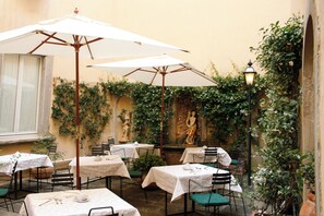 An outdoor café with tables and chairs, covered by umbrellas, and surrounded by greenery and a statue. - B&B Piccolo Hotel (Cortona)