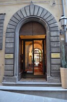 A hotel bejárata íves kapuval, egy tábla, amelyen az „Albergo I Michele” felirat látható, és a jobb oldalon egy virágtartóban lévő növény.