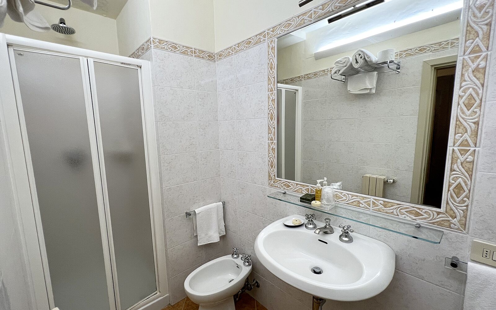 A bathroom with a glass shower enclosure, a white sink with a glass shelf, and a large mirror with decorative tiles.