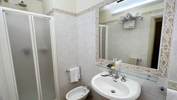 Un baño con cabina de ducha de vidrio, lavamanos blanco con repisa de vidrio y un espejo grande con detalles de azulejos decorativos.