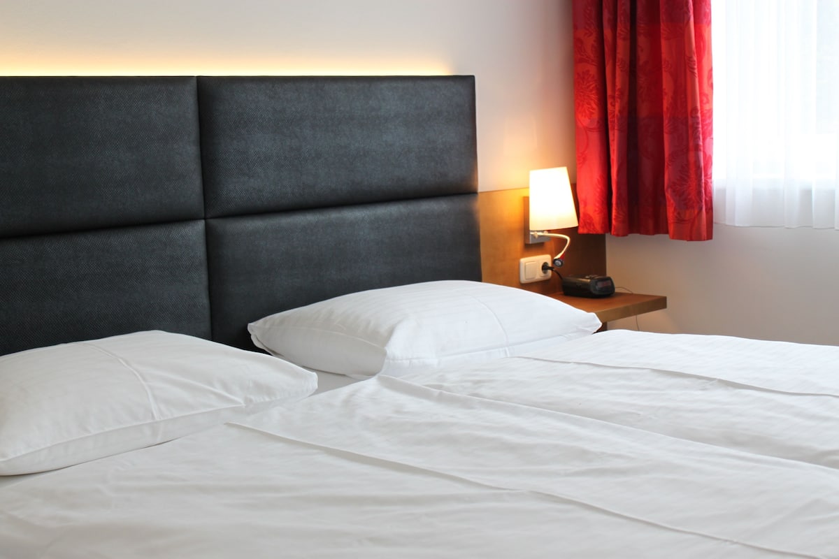 A hotel room with a large bed, a nightstand with a lamp, and a window with red curtains.