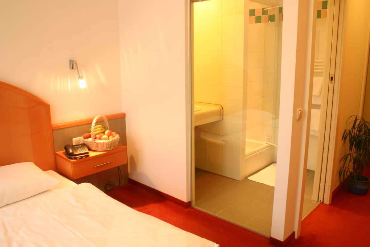 A hotel room with a bed, a bedside table with a basket of fruit, a telephone, and a glass-enclosed shower area.