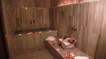 A spa room with a wooden bench, candles, and a small table with a towel and a copper bowl.
