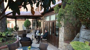 Un patio al aire libre con mobiliario de mimbre, una mesa de vidrio, paredes de piedra y abundante vegetación.