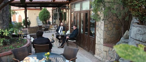 Un patio al aire libre con mobiliario de mimbre, una mesa de vidrio, paredes de piedra y abundante vegetación.