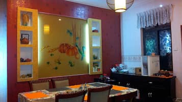 A dining area with a table set for two, chairs, a decorative wall with shelves, a hanging light fixture, and a kitchen area with appliances.