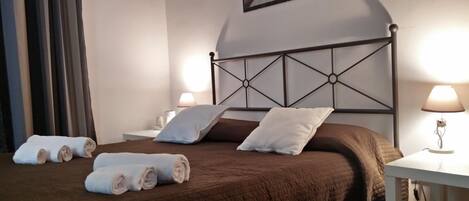 Una cama bien tendida con una colcha marrón, dos almohadas y toallas enrolladas sobre un piso de madera.