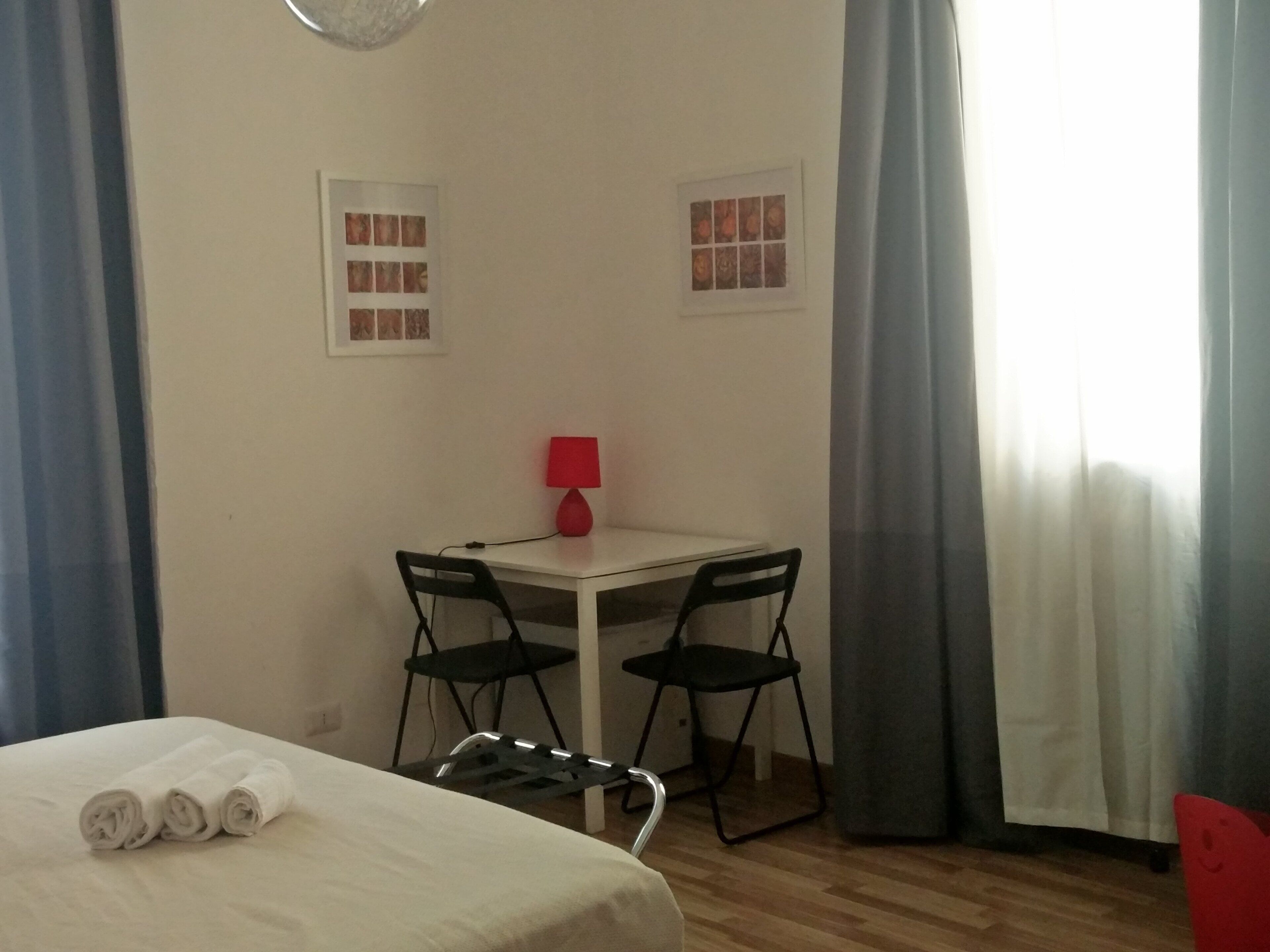Una habitación pequeña con una cama, una mesa con sillas, una lámpara roja y dos cuadros en la pared.