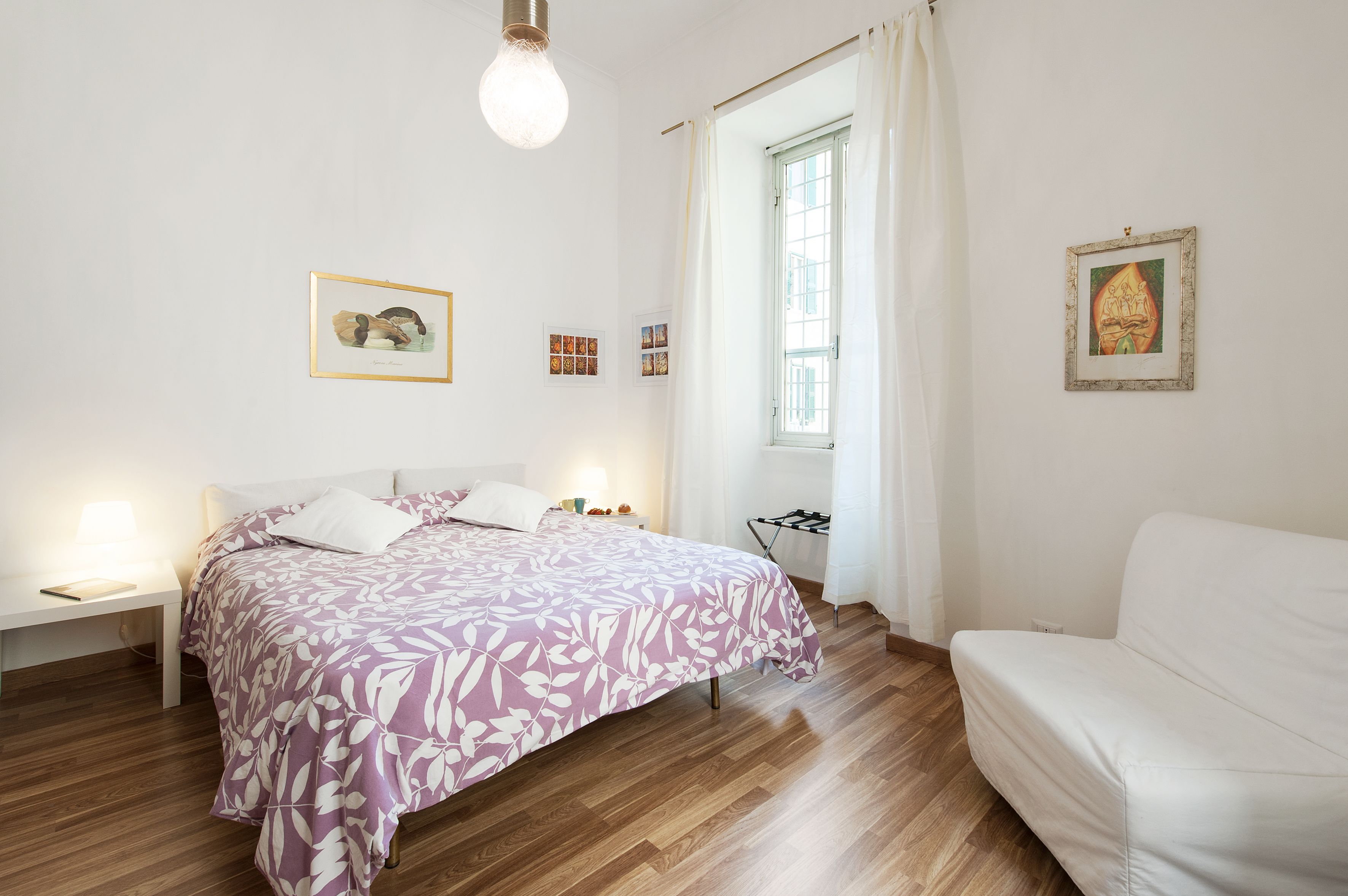 Un dormitorio con una cama, mesitas de noche, una silla blanca y obras de arte en las paredes.