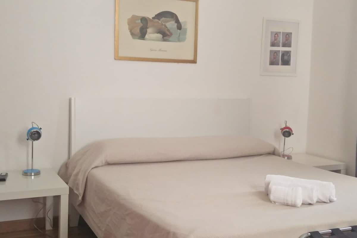 A neatly made bed with a beige blanket, a framed picture of birds on the wall, and a small table with a lamp beside it.