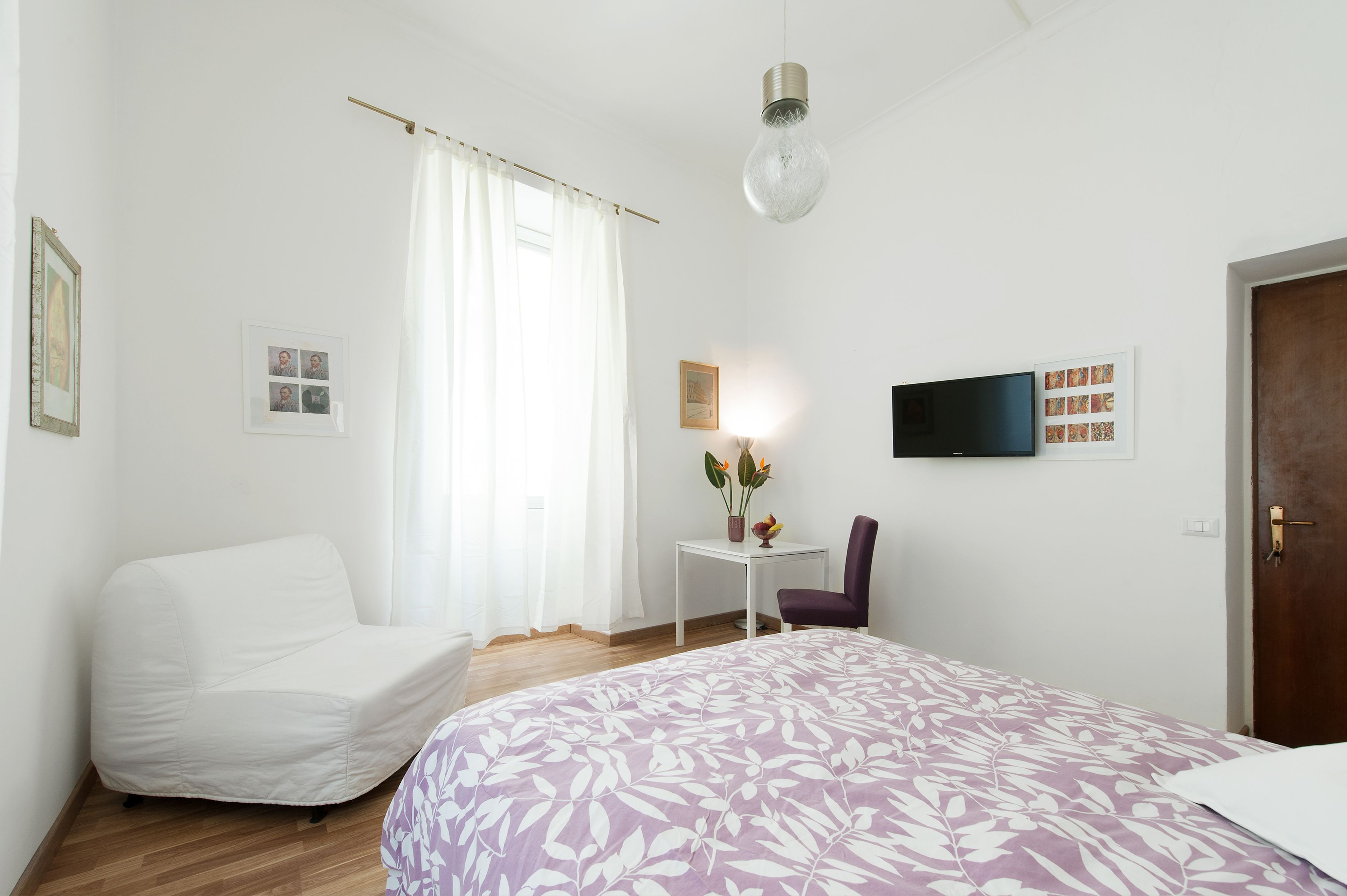 Un dormitorio con cama, una silla, una mesita con flores, un televisor y una puerta.