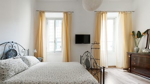 Ein Schlafzimmer mit Bett, Fernseher, Kommode und zwei Fenstern mit Vorhängen.