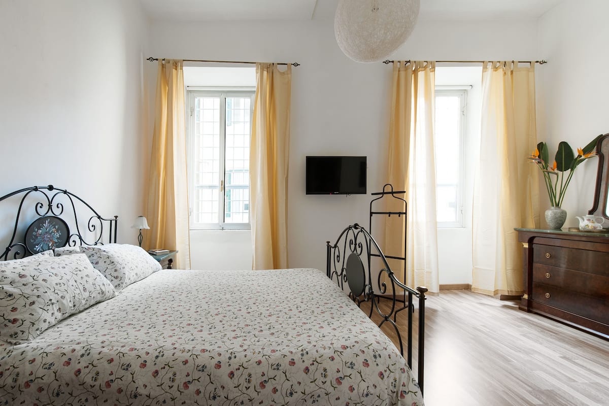 A bedroom with a bed, a TV, a dresser, and two windows with curtains.