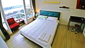 A hotel room with a large bed, a red chair, a desk, and a view of a cityscape through the window. - Impact Novo Condo Muang Thong Thani (Pak Kret)