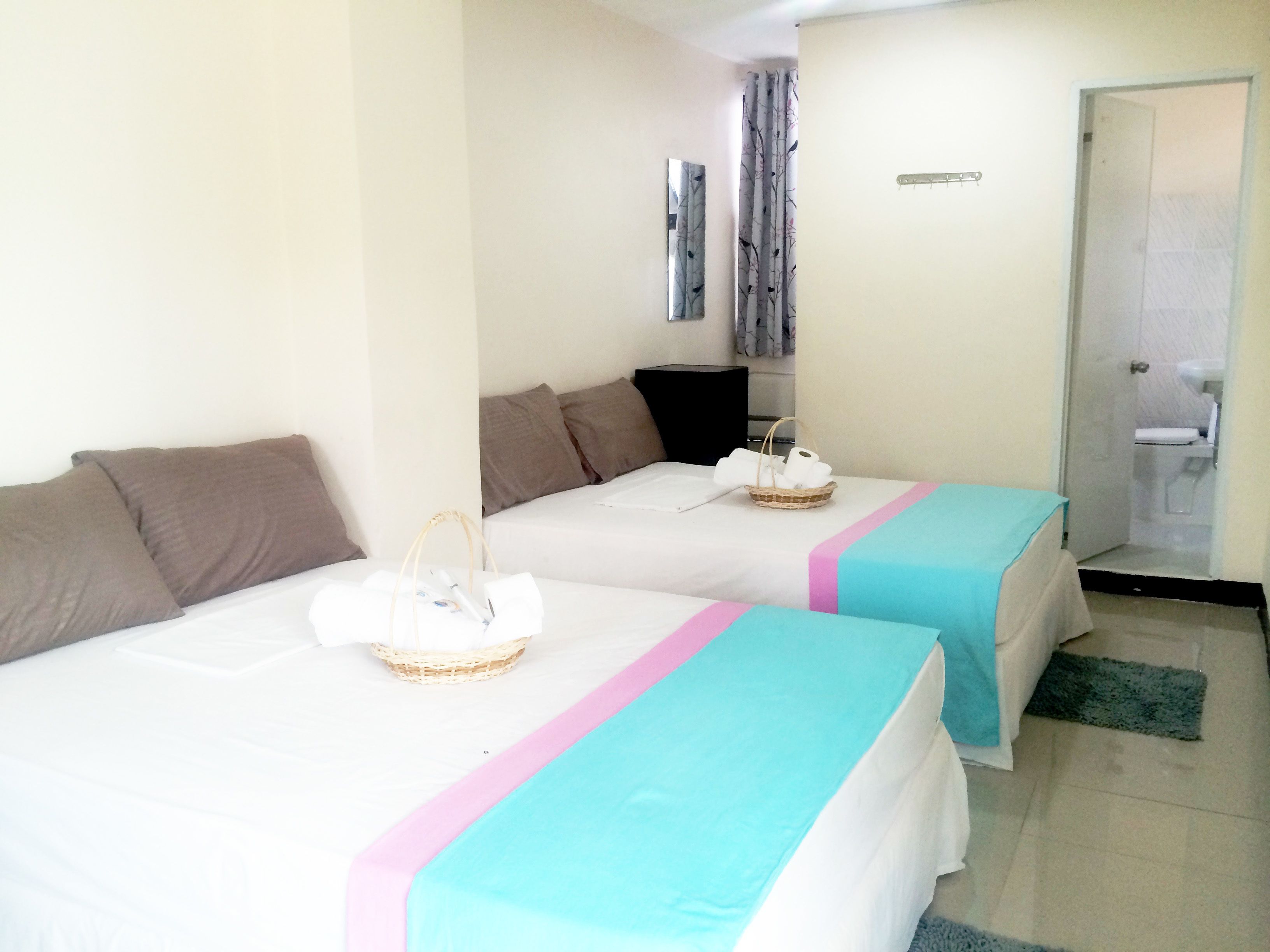Una habitación de hotel con dos camas, cada una con una colcha verde azulada y rosa, un cesto de artículos de tocador y un estante pequeño sujeto a la pared.