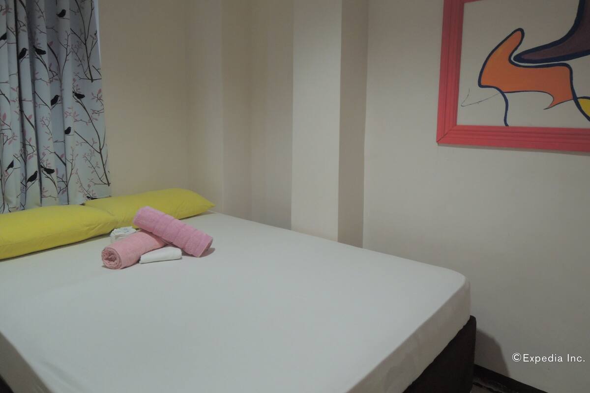 Una cama individual con colchón blanco, dos almohadas (una amarilla y una rosa), y una toalla rosa enrollada. En la pared a la derecha, hay una obra de arte abstracta enmarco.