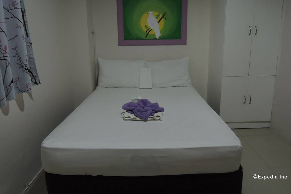 Habitación de hotel con cama, una toalla morada, una obra de arte enmarcada de un pájaro sobre una rama y un armario blanco.