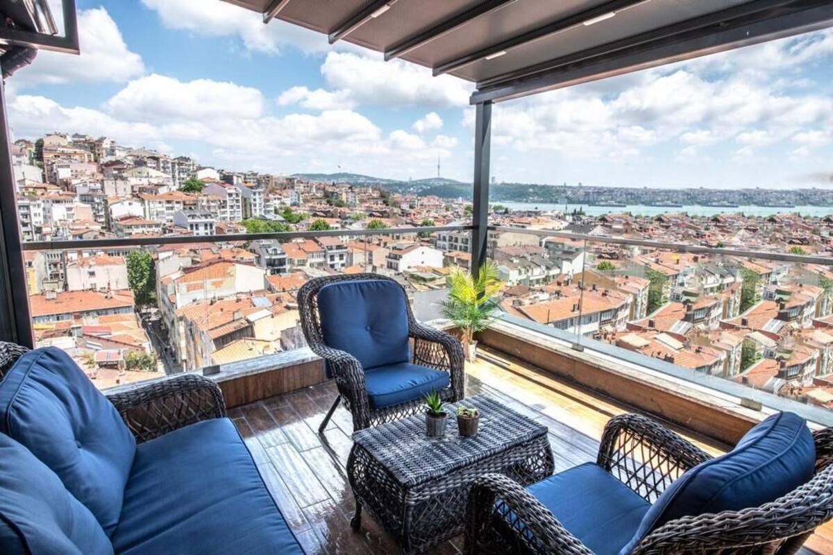 Un balcon agrémenté de meubles en osier, d’une plante en pot et offrant une vue sur un paysage urbain dense.