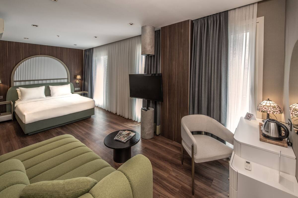Une chambre d’hôtel moderne avec un grand lit, un téléviseur à écran plat, un canapé vert, un fauteuil blanc et une petite table avec un magazine.