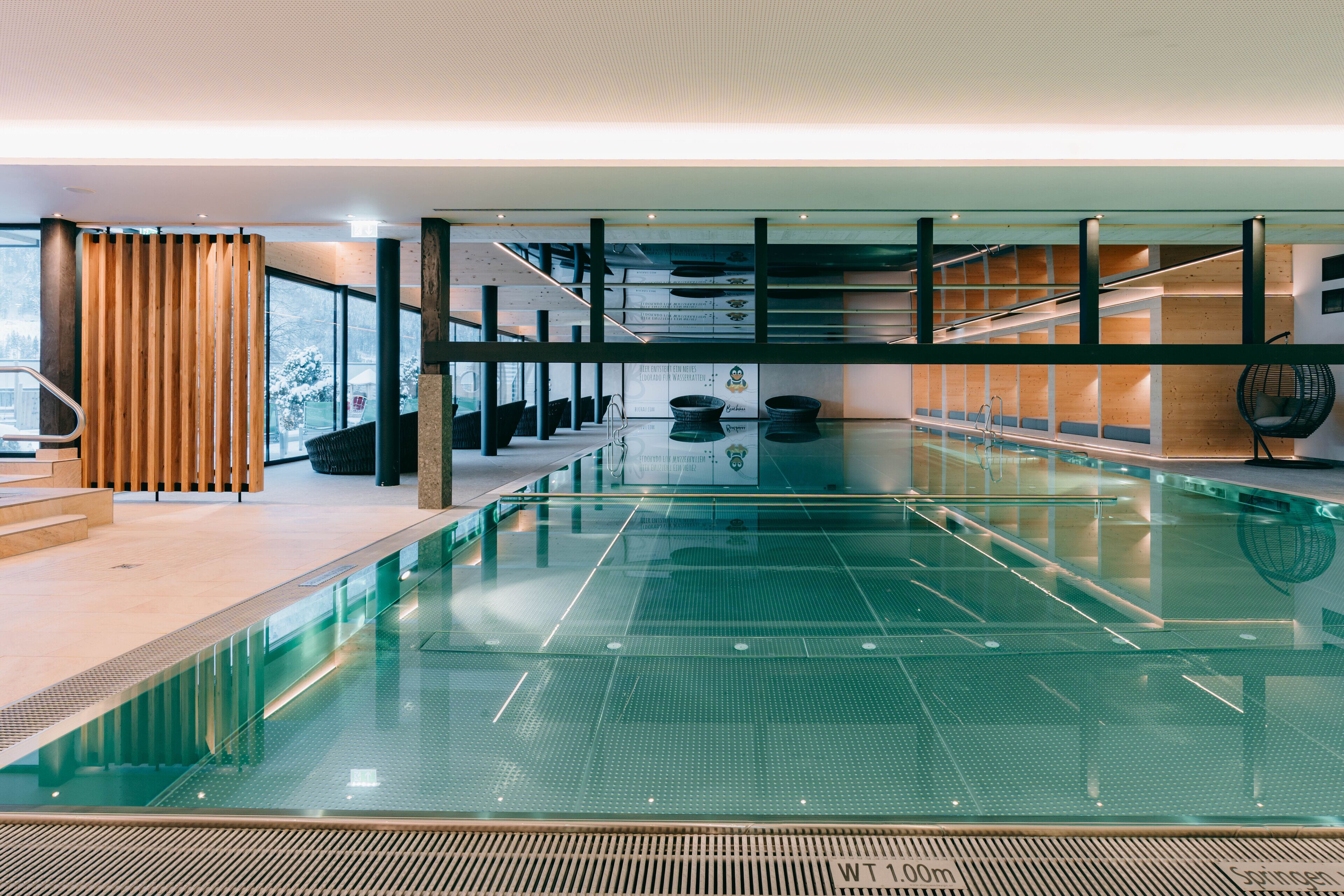 Una piscina interior moderna con paredes de vidrio transparente, paneles de madera y un indicador de profundidad visible.