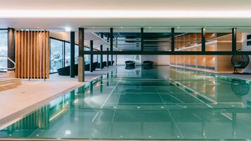 Una piscina interior moderna con paredes de vidrio transparente, paneles de madera y un indicador de profundidad visible.