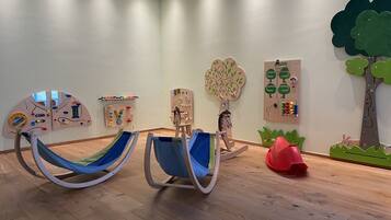 Una habitación con dos mecedoras, un espacio de juego de madera con juguetes educativos y una pared decorada con un árbol y un conejo.