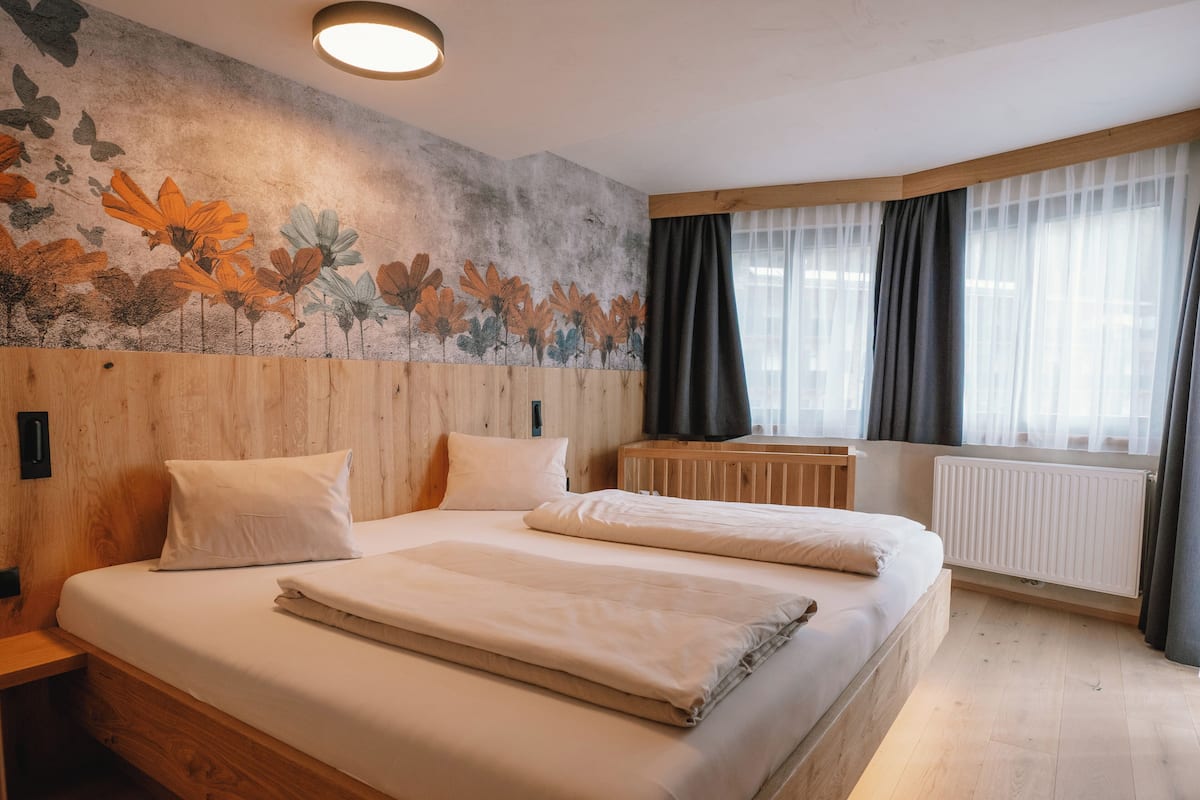 Une chambre avec une tête de lit en bois, un grand lit avec une literie blanche, une fresque florale sur le mur et une fenêtre avec des rideaux.