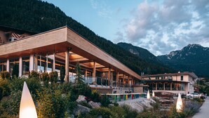 Un bâtiment moderne doté d’une grande avancée et de nombreuses fenêtres, entouré d’une végétation luxuriante et avec des montagnes en arrière-plan.